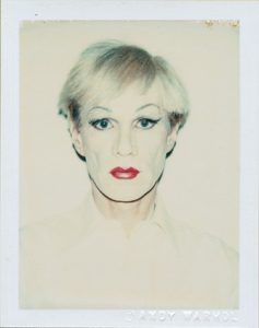 A photograph of Andy Warhol dressed in drag. He wears white face paint, winged eyeliner, and bright red lipstick. He has on a white shirt, which almost blends in with the pale background.