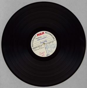 A vinyl record sits against a gray background. On the paper in its center is David Bowie's signature in blue ink.