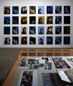 A grid of Warhol's Jackie portraits silkscreened in blue, yellow, and black, with a vitrine in front filled with Jackie memorabilia.