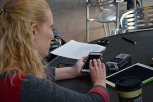 A young woman with blonde hair is seated at a table. She holds an old iPhone, which she is using to test out the new audio guide app.