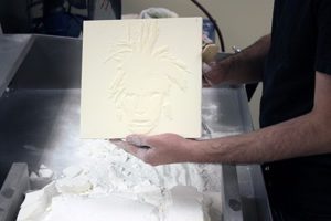 A man holds a while plastic tile. On the tile, a self portrait of Andy Warhol has been recreated in 3D, which will allow museum patrons to experience the art in a new, tactile way.