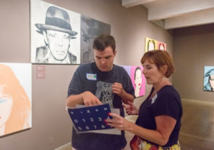 A young man and a women stand in a gallery filled with colorful celebrity pop portraits. The young man in pointing at a sheet of paper the women is holding.