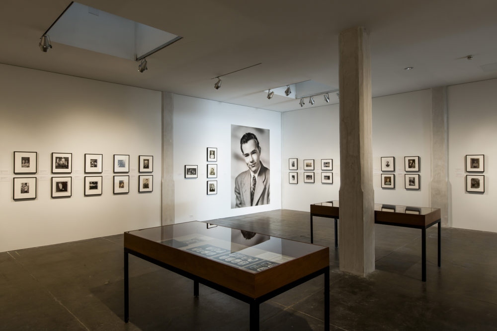 A large photograph of Andy Warhol from high school is in the center surrounded by smaller framed black and white photographs. In front on the photographs are two display cases housing pages from photo albums full of small black and white photographs.