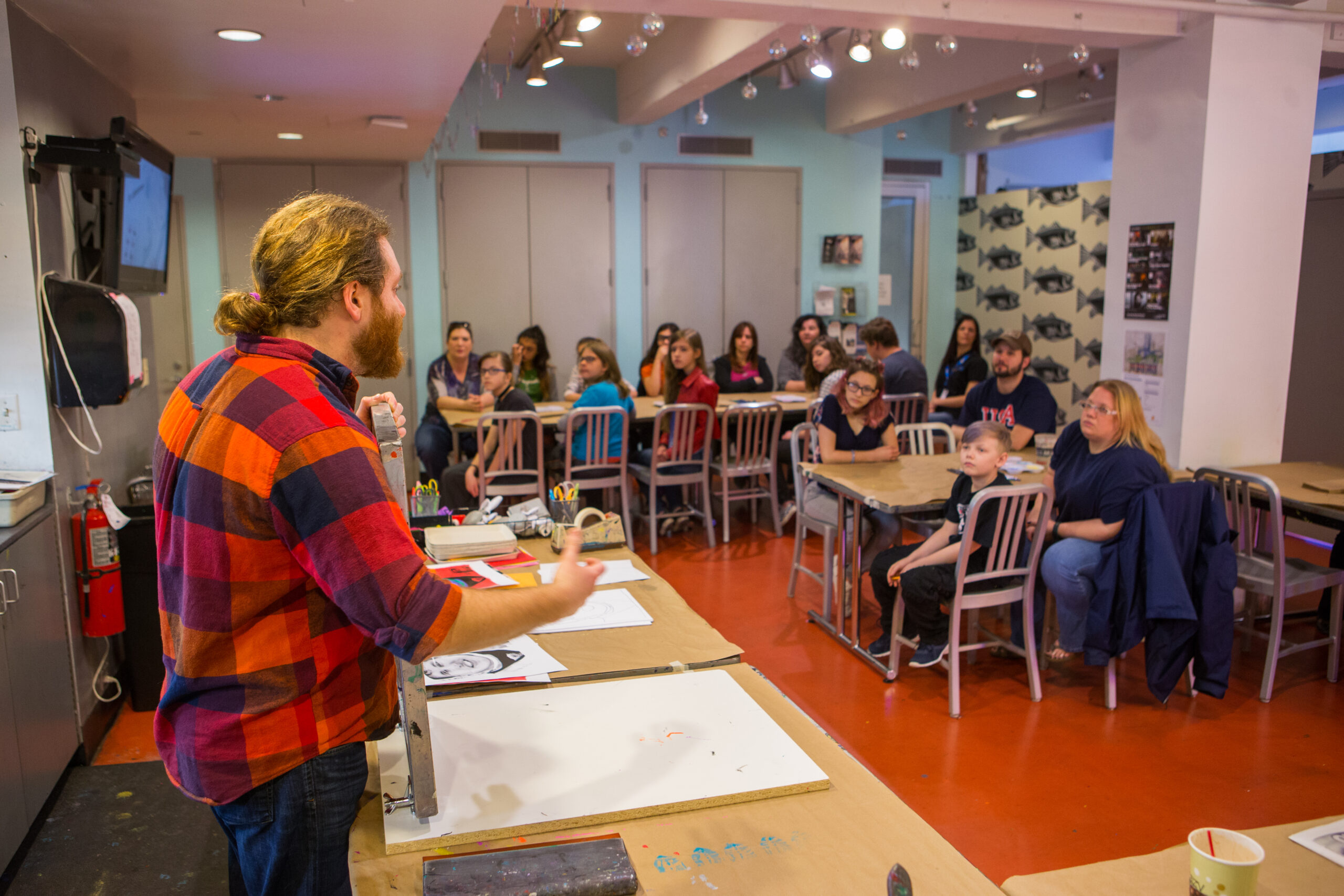 A male artist educator with long blond hair pulled back into a bun wearing a red plaid shirt presents information to students sitting in silver chairs at long, rectangular tables in the Andy Warhol Museum’s studio.