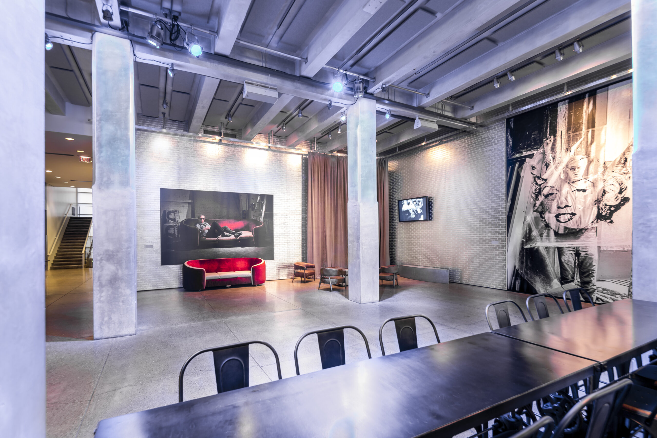 A photograph of the Andy Warhol museum entrance space. There is a long, silver table in the foreground and a red velvet couch pushed up against a far wall under a portrait of Andy Warhol reclining on a red velvet couch.