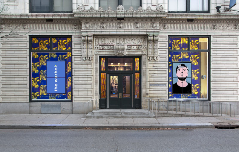 A photograph of the Andy Warhol Museum entrance. It is an ornate building with cement stairs and an accessible ramp that leads off the right side of the image. A portrait of Marilyn Monroe is visible through glass panels in and around the front doors. The front door is flanked by two large windows with blue wallpaper featuring screen prints of yellow cows. The window on the left side of the image has a sign that reads The Warhol in white print. The right window has a screen-printed portrait of Andy Warhol.