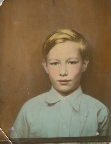 This faded photograph depicts school-aged Andy Warhol wearing a blue button-up shirt standing against a brown background, his blonde hair neatly combed.