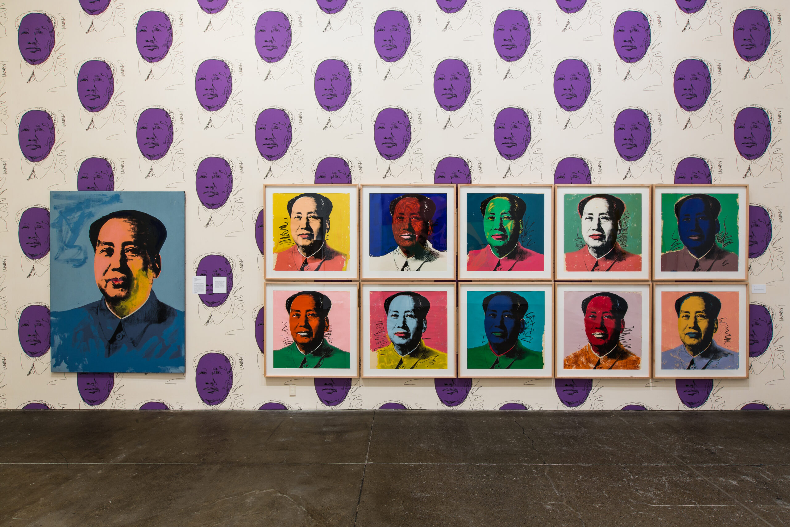 Eleven Mao portraits in different colors hanging on a wall wallpapered with purple Mao portraits.