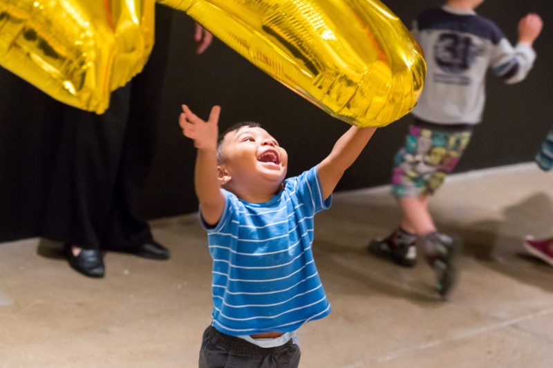 A smiling child throws a gold balloon up in the air in a gallery