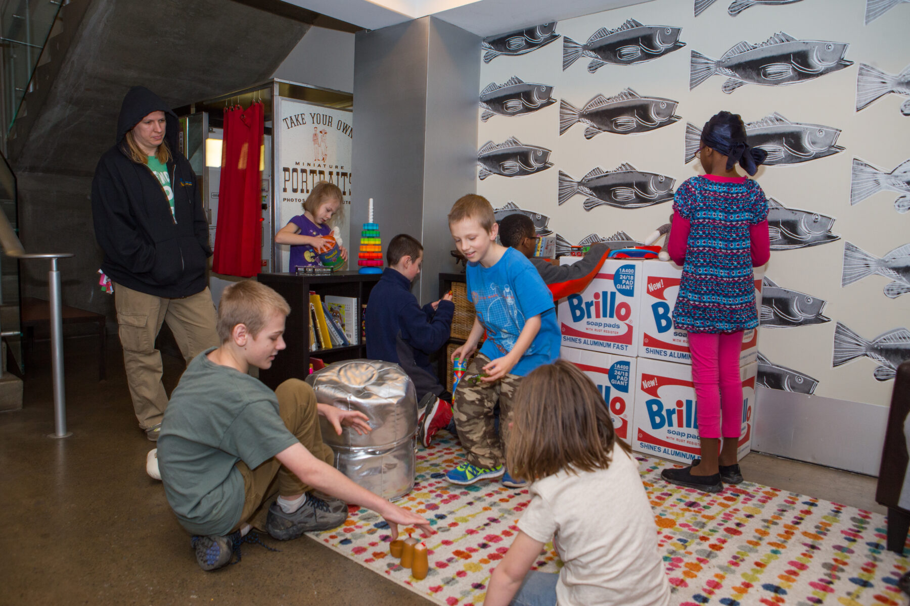 A group of children plays with toys in the Andy Warhol Museum's underground level as a woman in a black hoodie looks on.