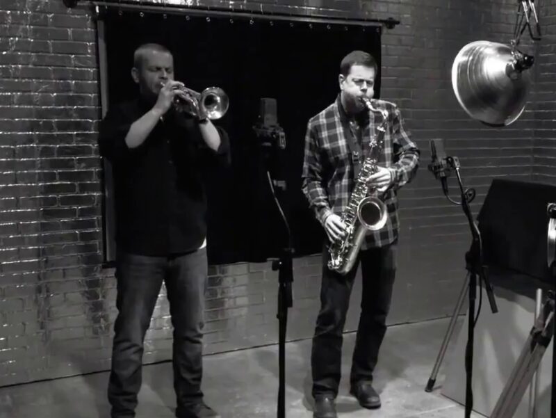 Screen grab from live performance, in black and silver. Two men stand in-front of the Andy Warhol Screen Test machine, playing the saxophone and trumpet.