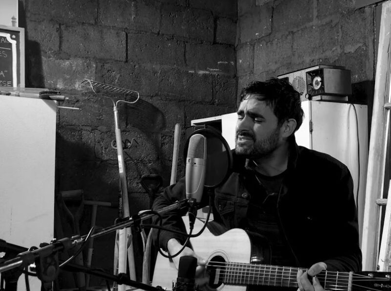 Screen grab from live performance, in black and silver. Andre Costello sings and plays the guitar.