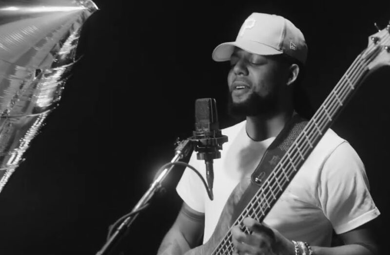 Screen grab from live performance, in black and silver. Benji sings and plays the guitar in front of a microphone. On the left, one of Andy Warhol's Silver Cloud is partially visable