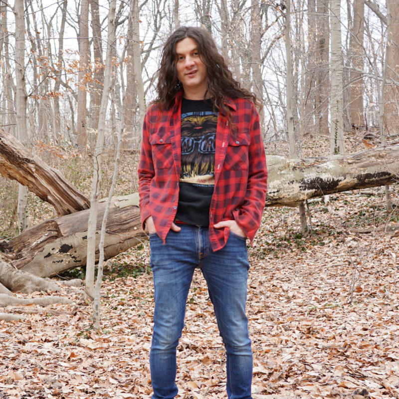 Man with long hair stands in a clearing in the woods, hands in his pockets.