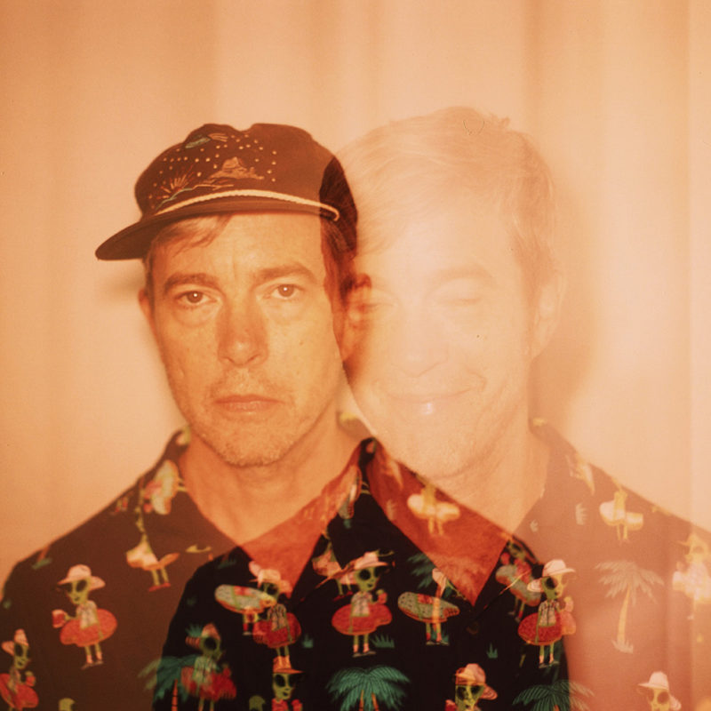 Double exposed photo of Bill Callahan in a button down shirt with vacationing aliens and palm trees on it. In one of the exposures he is wearing a hat and looking stoically at the camera. In the other exposure, he is not wearing a hat and is smiling.