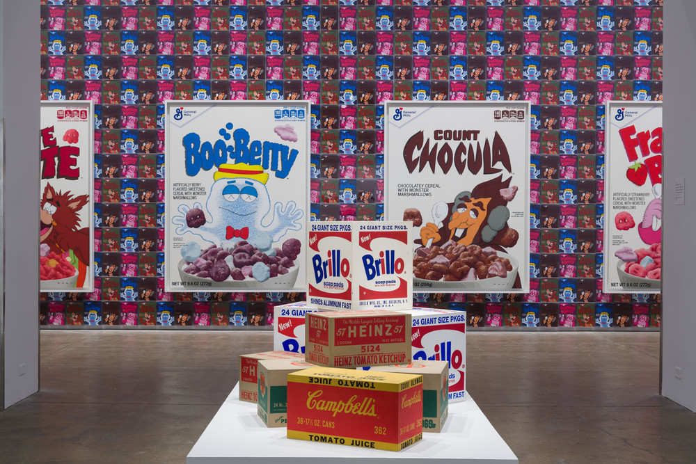 Box sculptures on a plinth in the foreground with two large paintings of cereal boxes in the background.