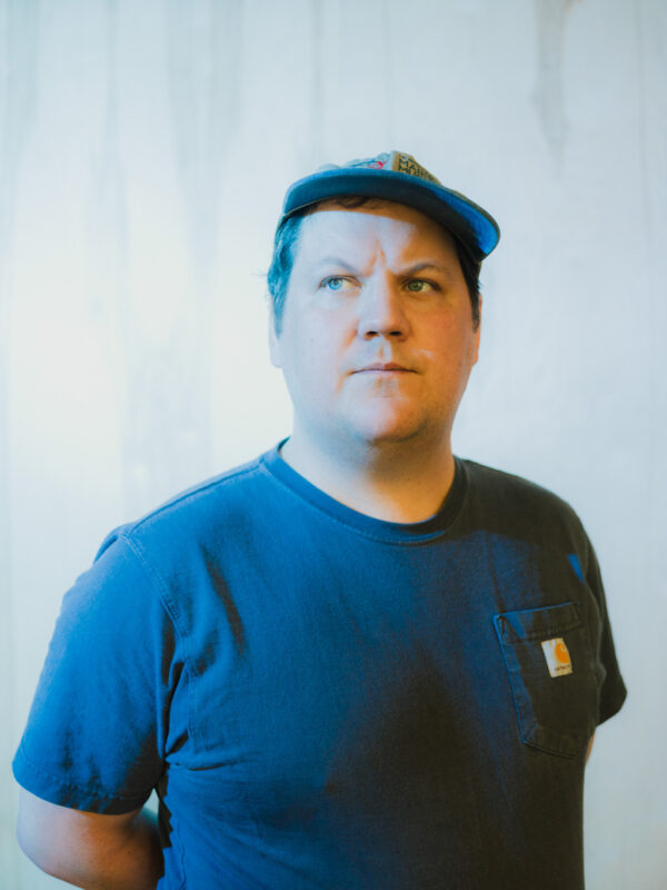 Person wearing a blue t-shirt and a baseball cap, standing against a light-colored backdrop.