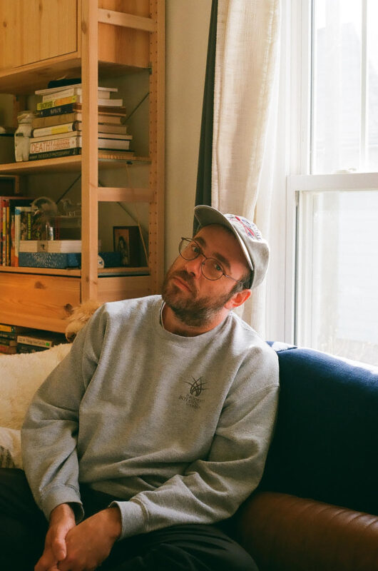 Person in a grey sweatshirt and cap sitting by a window with a bookshelf in the background.