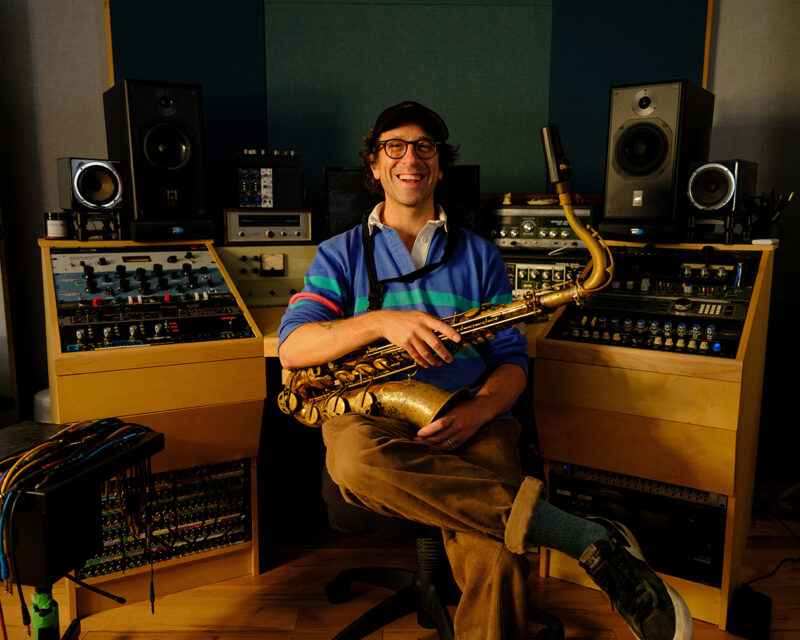 Person smiling, holding a saxophone, seated in front of a sound mixing board and speakers in a recording studio.