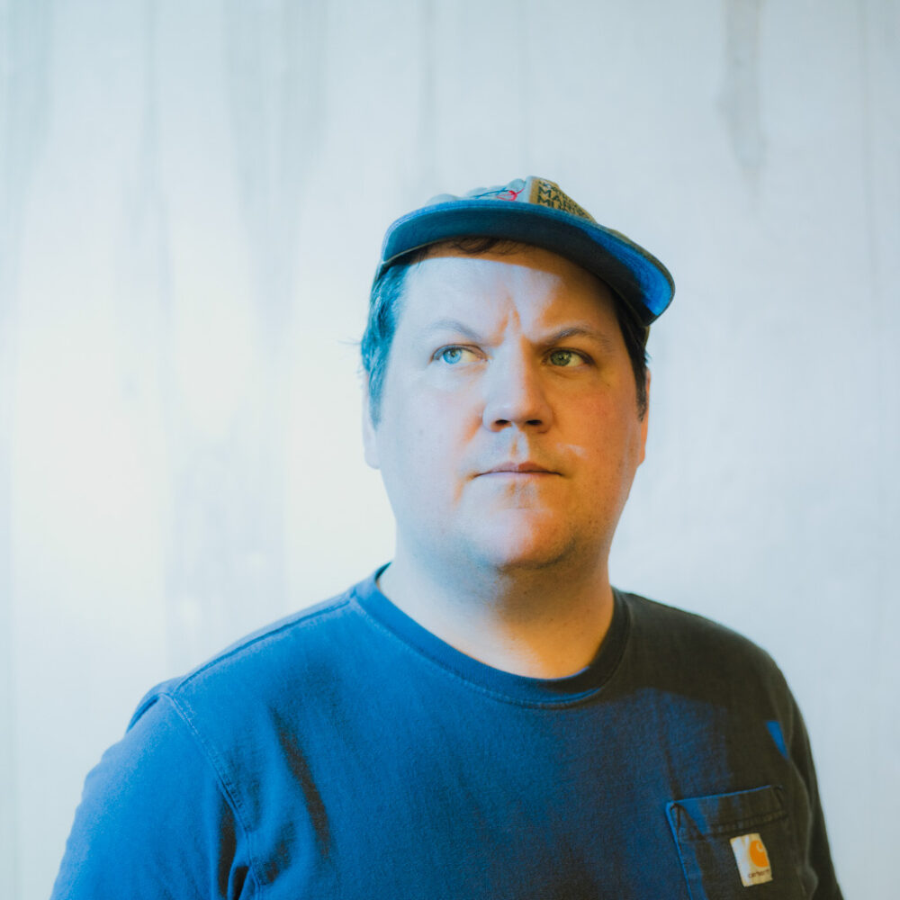 Person wearing a blue t-shirt and a baseball cap, standing against a light-colored backdrop.