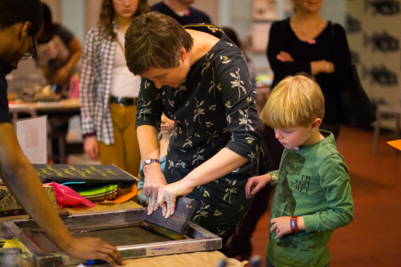 A person uses a squeegee to push ink through a silkscreen while a child watches.