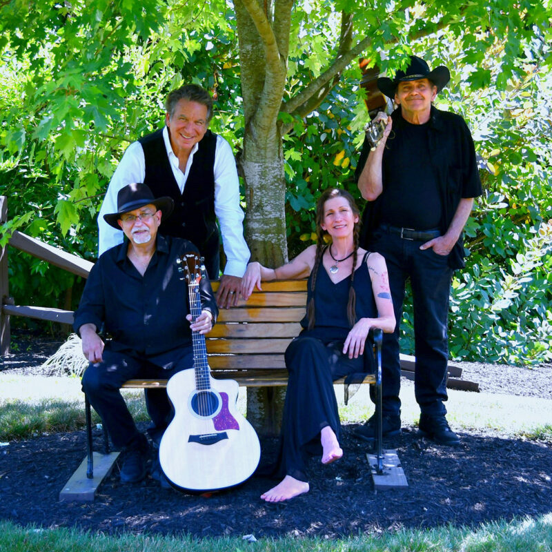 Four members of a band posing outdoors with a guitar, two sitting on a bench under a tree while two others stand behind the bench, all dressed in casual attire.