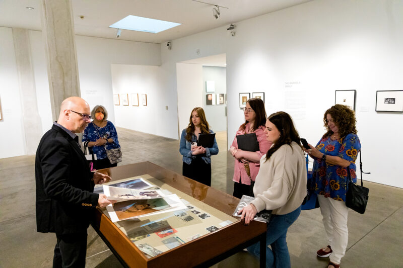 A man with a suit coat and glasses stands to the left of a vitrine in a gallery showing laminated images of Shirley Temple to a group of five teachers standing around the right side of the vitrine.