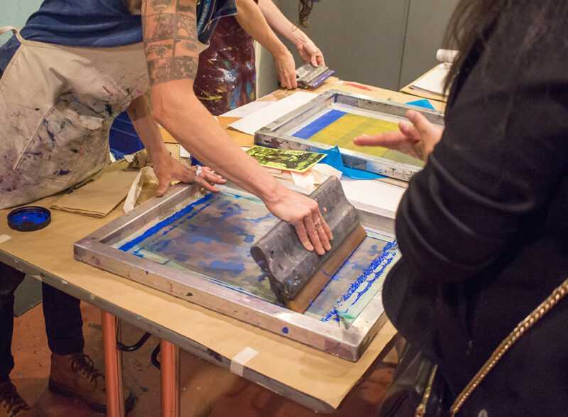 A close-up view of two people silkscreen printing a camoflauge pattern. The person on the left wears a paint-splattered apron and holding a squeegee covered in blue ink, while the person on the right wears a black hoodie and watches. In the background, a third person is preparing to silkscreen print.