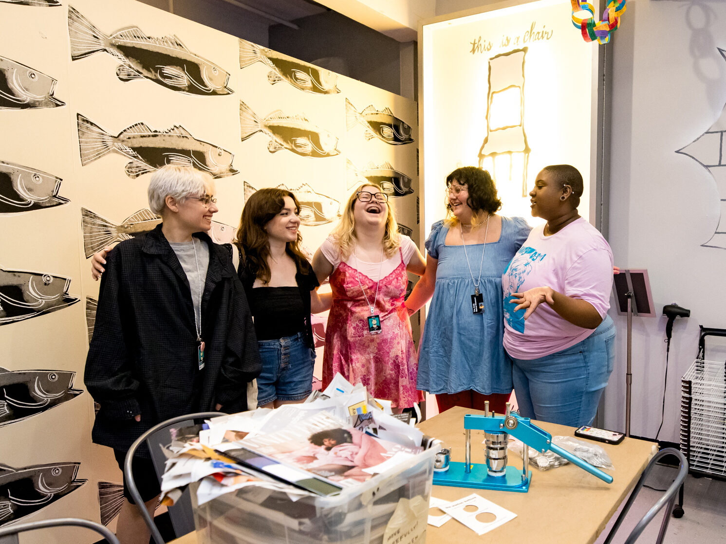 A group of five individuals standing in a room that appears to be an art studio or classroom, indicated by various artworks on the walls, including drawings of fish and abstract shapes. A colorful paper chain hangs from the ceiling. In the foreground, a table is cluttered with items such as papers, a plastic storage box, and what seem to be art supplies.