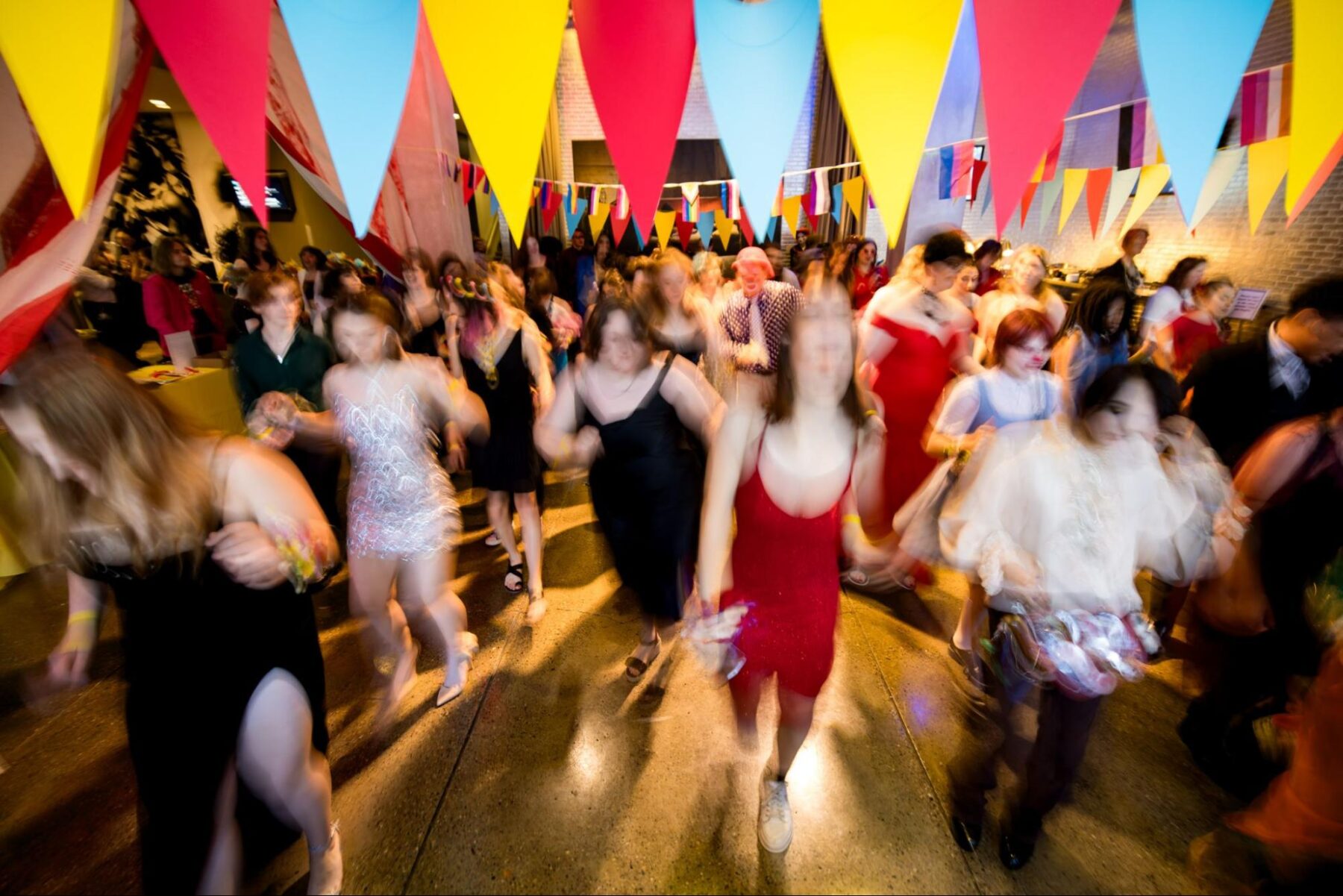 An evening event with many people moving around, possibly dancing or walking. Colorful triangular flags are strung above the crowd, adding to the festive atmosphere. The lighting is dim but warm, highlighting the energy and movement of the gathering.