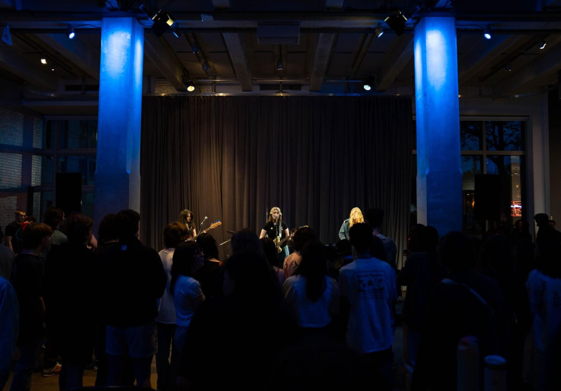 three musicians performing on stage in a dimly lit venue. One musician on the left is playing a guitar, while the two in the center and right are singing into microphones. The audience is standing and facing the stage, engaged with the performance. The stage has blue lighting accents and dark curtains in the background.