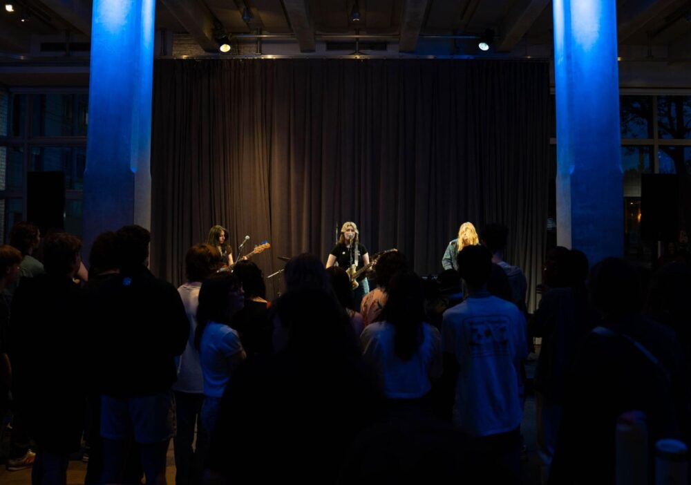 three musicians performing on stage in a dimly lit venue. One musician on the left is playing a guitar, while the two in the center and right are singing into microphones. The audience is standing and facing the stage, engaged with the performance. The stage has blue lighting accents and dark curtains in the background.