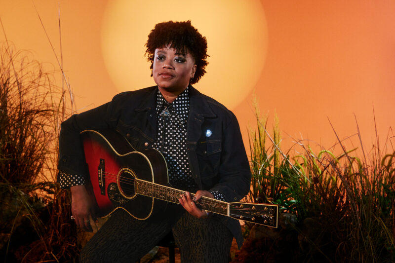 Amythyst Kiah sitting with a guitar on her lap against an orange backdrop with silhouettes of tall grass and a sun behind her head.