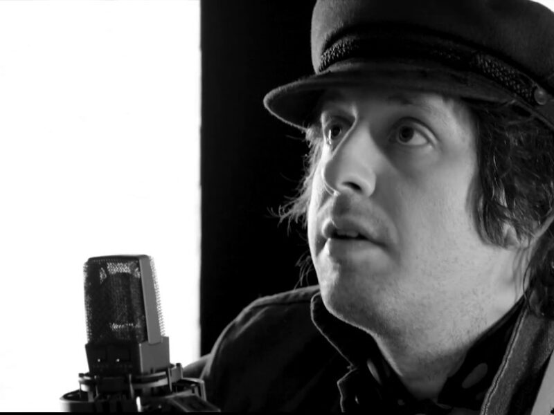 Screen grab from live performance, in black and silver. Adam Green sings into a microphone