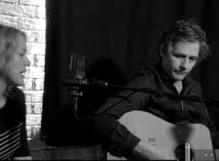 Screen grab from live performance, in black and silver. Dean plays a guitar in front of a mike in the center of the image, and Britta sings into a microphone on the left.