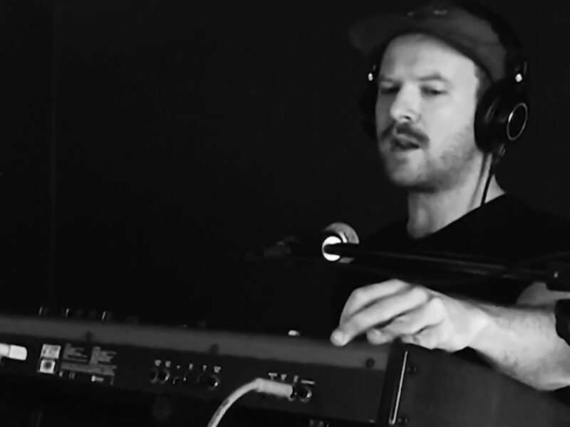 Screen grab from live performance, in black and silver. A member of Good Sport plays they keyboard.