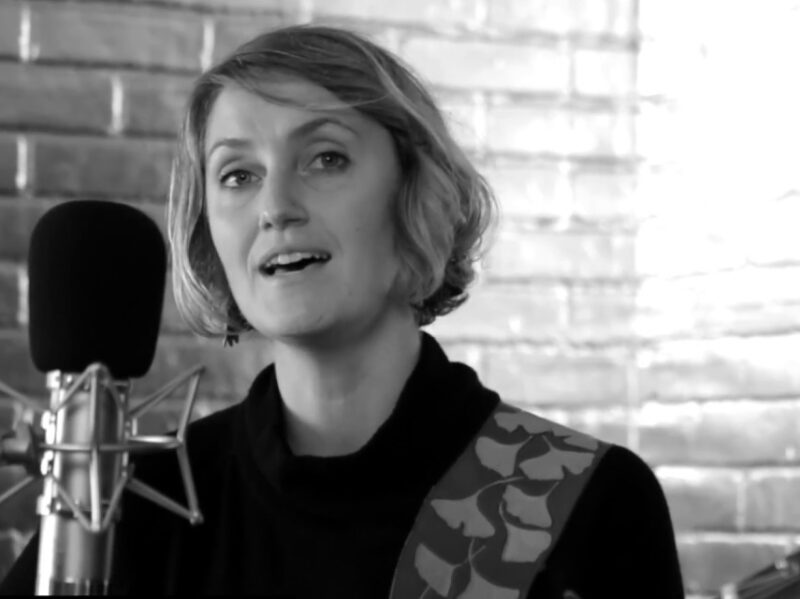 Screen grab from live performance, in black and silver. Joan Shelley sings into a microphone while holding a guitar.