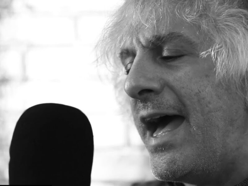 Screen grab from live performance, in black and silver. A close up of Lee Ranaldo singing into a microphone.