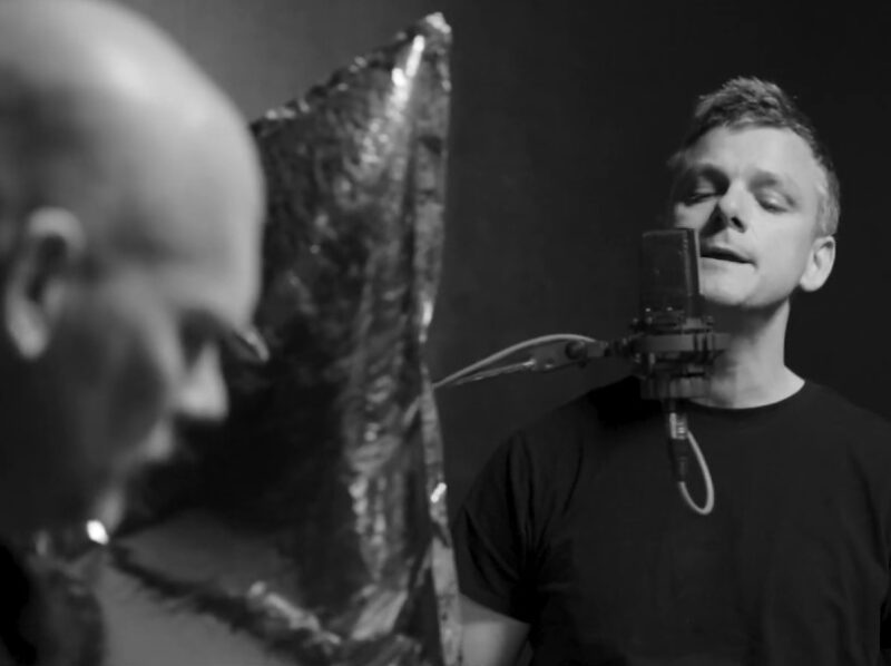 Screen grab from live performance, in black and silver. The lead singer sings into a microphone, while the guitarist plays his guitar in the foreground.