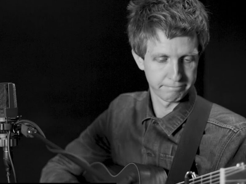 Screen grab from live performance, in black and silver. A musician looks down while playing the guitar