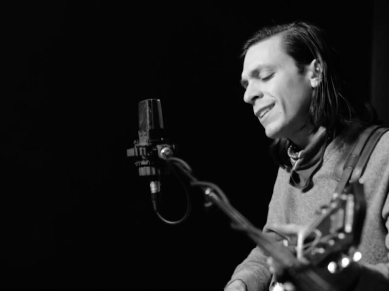 Screen grab from live performance, in black and silver. Anthony LaMarca plays his guitar and sings.