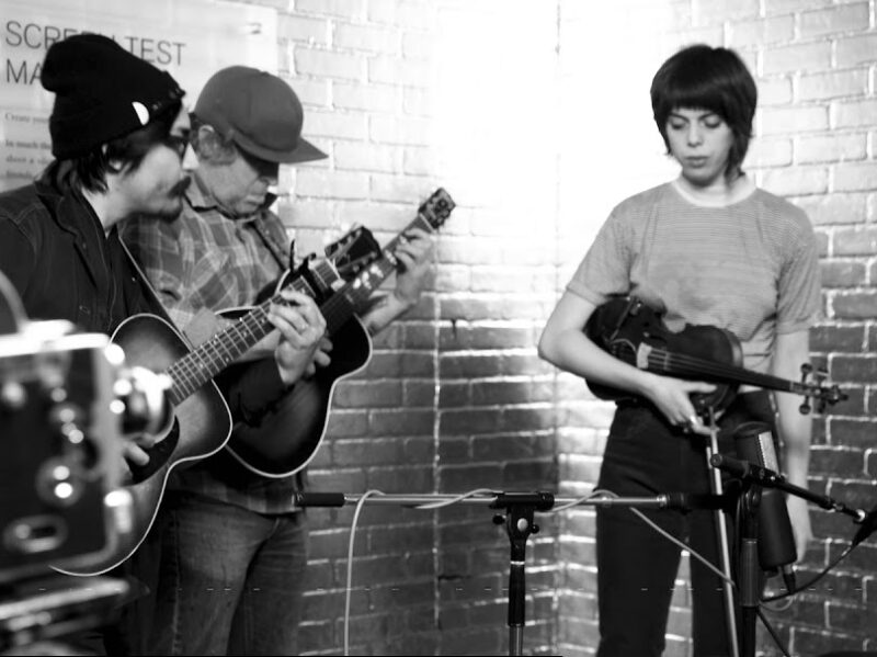 Screen grab from live performance, in black and silver. Four members of The Low Anthem play their instrument. One, the singer, is sitting.