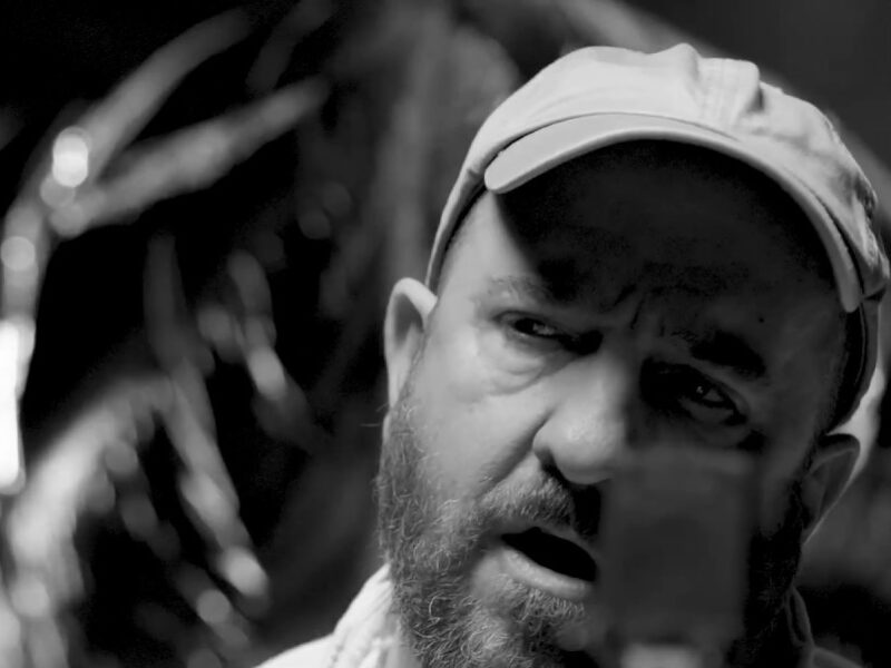 Screen grab from live performance, in black and silver. A bearded man with a hat sings into a microphone,