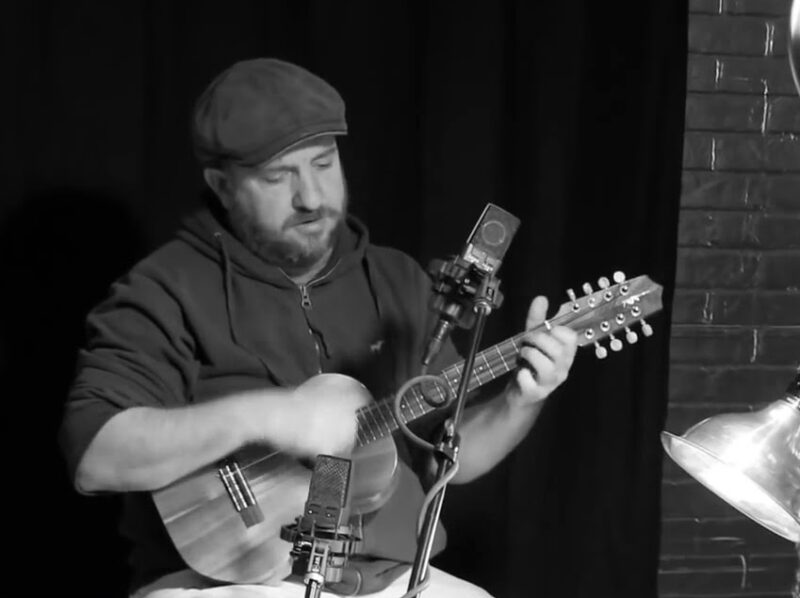 Screen grab from live performance, in black and silver. A man sings into a microphone, and plays a guitar.