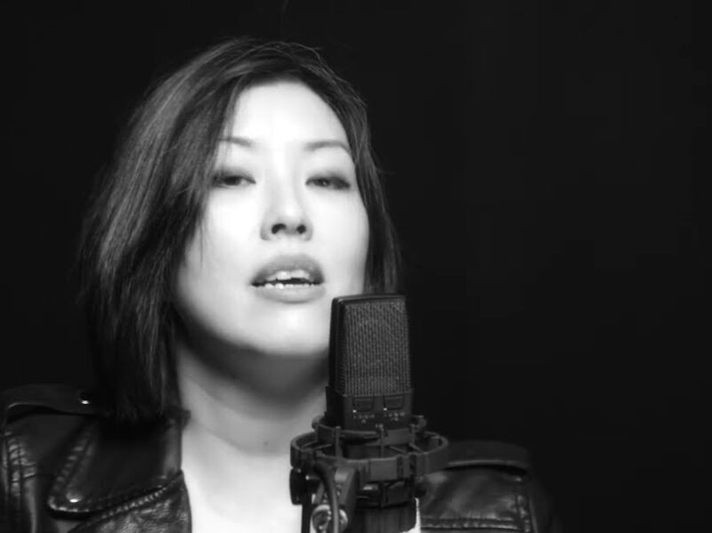 Screen grab from live performance, in black and silver. Sharon Mok sings into a microphone with a black background.