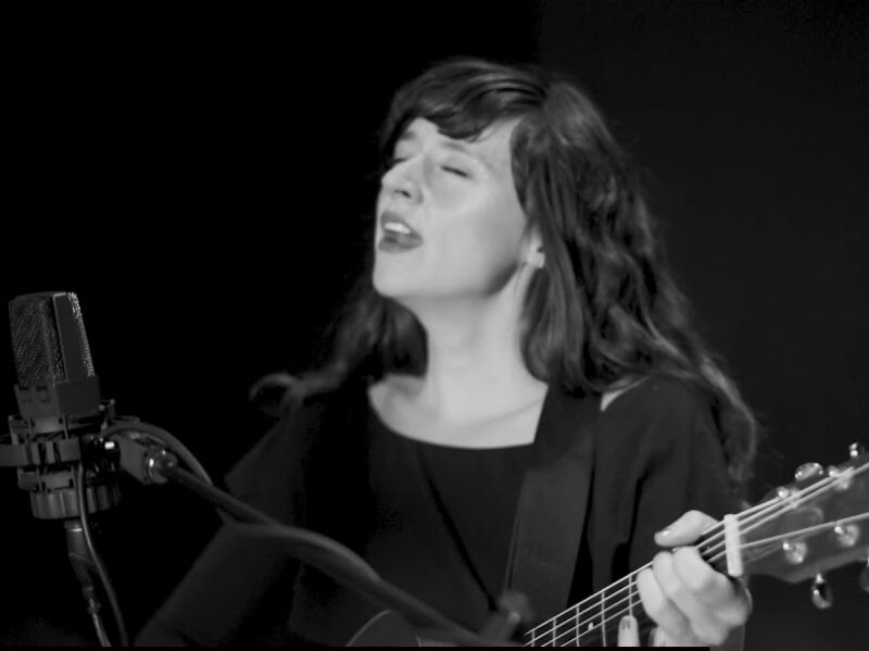 Screen grab from live performance, in black and silver. The artist sings and plays their guitar