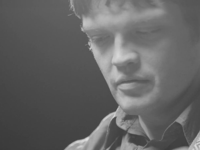 Screen grab from live performance, in black and silver. A close up of William Tyler's face, looking down at his guitar.