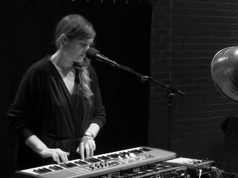 A black and white screen grab woman plays an electric keyboard and stands in front of a microphone.