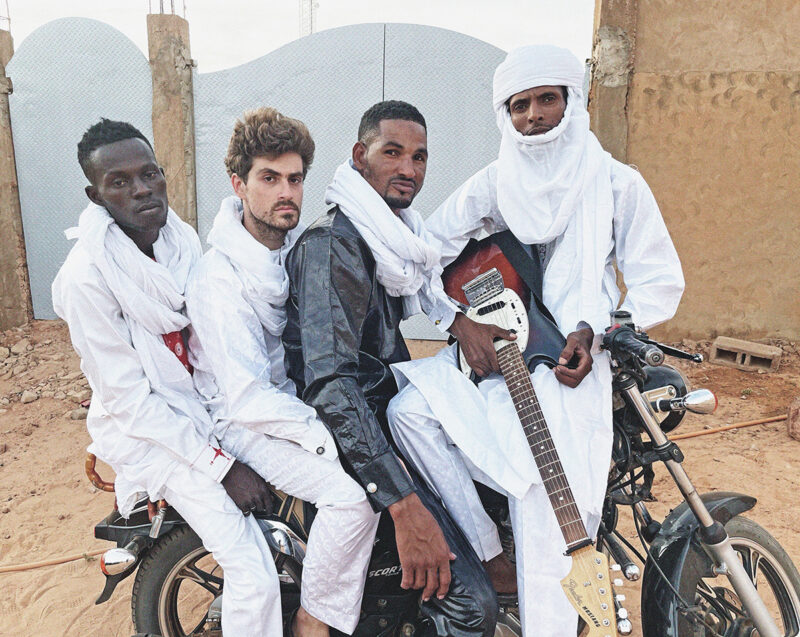 Mdou Moctar and three members of his band are dressed in traditional Tuareg attire, posing with a motorcycle in a desert setting. One member holds an electric guitar.