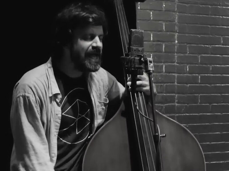 A black and white screen grab of a man standing and playing an upright bass while singing into a microphone.